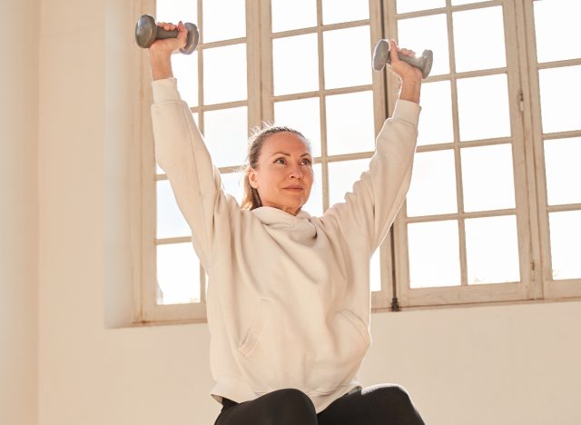 mature woman doing the overhead press with dumbbells, concept of strength exercises to keep fit in your 50s