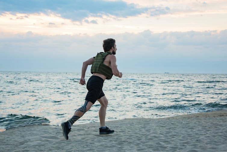 A young man wearing a weighted vest runs along a beach.